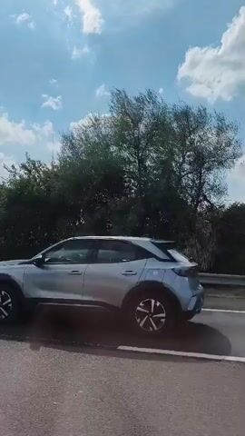 The video begins with a silver car driving on a road, the camera is looking out the window. A windmill is visible in the background to the left of the frame, partially obscured by trees. The car passes a stretch of trees and bushes along the roadside. The windmill becomes more prominent and then disappears behind trees again. The video then shows a large white truck passing by, its wheels and lower body are visible. The truck is on the same road.