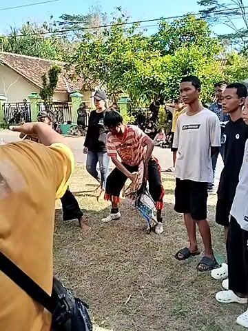 The video shows a gathering of people outdoors, possibly in a rural area, with trees and houses visible in the background. Several individuals are dressed in traditional clothing, some appearing to be engaged in a performance or demonstration. One person in the foreground, seen from behind, wears a yellow shirt and a black crossbody bag, with blue-tinted hair styled with dreadlocks. In the middle ground, two individuals in black shirts and jeans are holding up phones, appearing to record. Another person, in a striped shirt and black pants with red trim, is holding a prop that resembles a large bird. Several other people are scattered around, some watching the activity, others participating. The ground is covered in dry grass, and the overall lighting suggests it is daytime with a bright sun. Some of the participants in black uniforms are on the ground, seemingly engaged in a physical demonstration or fight. The scene suggests a cultural event or festival.