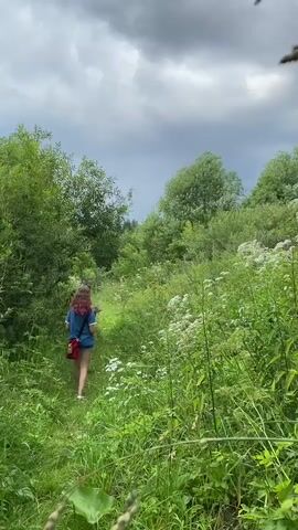A young person with long, curly red hair walks along a narrow, overgrown path through a lush green forest under a cloudy sky. They are wearing a blue t-shirt, denim shorts, and white sneakers, and carrying a red bag over their shoulder. The path is lined with tall green grass and white wildflowers, with trees and bushes on either side. The sky is filled with dark, ominous clouds.