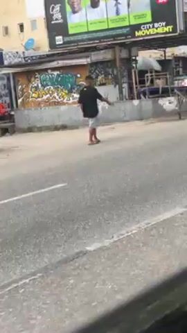The video shows a man standing in the middle of a street with his arm outstretched. He is wearing a dark shirt and light-colored shorts, and appears to be gesturing, possibly flagging down traffic. Behind him is a low concrete barrier covered in graffiti. Above the barrier is a billboard with several faces and the words "CITY BOY MOVEMENT." The road surface is asphalt with visible lane markings. The video is shot from a moving vehicle.