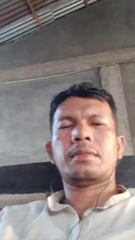 A man with dark hair and brown eyes, wearing a white collared shirt and earbuds, is facing the camera. He is sitting in front of a brick wall with a concrete ceiling. He raises his right hand and waves at the camera. The video is filmed indoors.