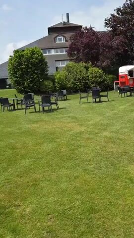 The scene shows a green lawn surrounded by buildings and trees. There are several black tables and chairs arranged in small groups on the lawn. Two red and white food trucks are parked on the lawn, with the words "Letz Spritz" visible on one of them. The buildings are modern and have large windows. The sky is blue with some clouds. The overall impression is that of an outdoor event or gathering space.
