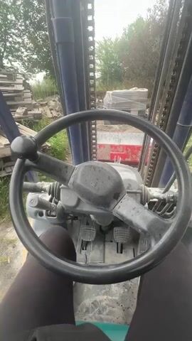 The video, set in Uhry, Ukraine, shows a first-person perspective from the driver's seat of a forklift. The camera captures the steering wheel, foot pedals, and the forklift's mast. Outside the forklift, on the left, stacks of wooden pallets are visible. On the right, a person is seen shoveling material, possibly sand or gravel, into a red wheelbarrow positioned in front of the forklift's forks. The scene appears to be an outdoor construction or storage yard with unpaved ground and scattered building materials. The weather is overcast. The forklift's forks then engage with the wheelbarrow, lifting it.