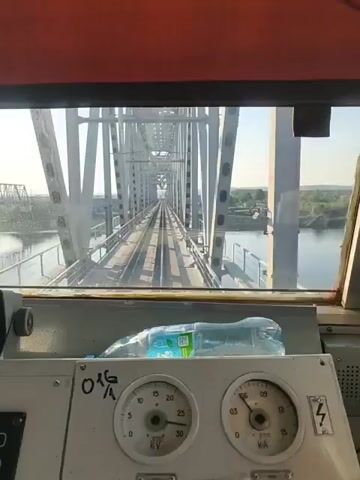 The view from inside the cabin of a train as it crosses a steel truss bridge is shown in the video. The bridge spans a wide body of water, with green vegetation visible on the distant banks. The train tracks extend straight ahead, framed by the bridge's structural elements. Inside the cabin, a dashboard is visible, featuring gauges and controls, with a plastic water bottle resting on top. The interior has a reddish-brown ceiling and a gray control panel. The day appears sunny and clear.