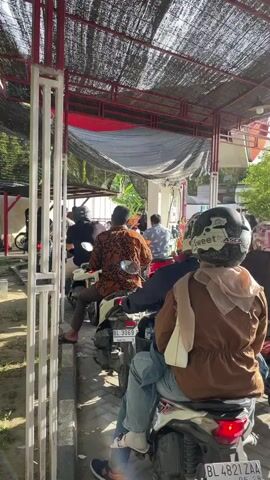 A group of people on motorcycles are lined up, waiting at what appears to be a gas station. The people are mostly facing away from the camera, with their backs visible. They are wearing helmets, some with designs. The motorcycles are parked in a row under a covered structure, with a black tarp overhead. The ground is paved with interlocking bricks. The lighting suggests it is daytime, with sunlight filtering through the covering. The overall scene depicts a common, everyday occurrence of people queuing for fuel.