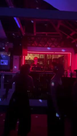 The video captures a lively scene inside a dimly lit nightclub or bar. The dominant lighting is neon pink and blue, creating a vibrant and energetic atmosphere. In the background, behind a bar with neon trim, a person wearing a red hat and headphones stands, presumably a DJ, overseeing the music. Several patrons are visible, silhouetted against the colorful lights, dancing and enjoying themselves. Their figures are dark, emphasizing the effect of the club's lighting.

The setting appears to be a tropical location, possibly a beach bar or open-air venue given the visible wooden beams in the ceiling and the presence of a ceiling fan on the left side of the frame. The presence of what looks like plastic cups on the bar further suggests a bar setting. The overall mood is one of celebration and enjoyment, with the music and dancing contributing to a sense of nightlife and revelry. There is no indication of the time of day other than it being nighttime due to the indoor lighting and the nature of the activity. No specific weather conditions can be inferred.

On the wall to the left of the DJ booth, there is a small sign with some text, but it is too blurry to be read. The scene is dynamic, with the camera seeming to be held by someone moving through the crowd, adding to the immersive feel of the experience. The activity is clearly a social gathering centered around music and dancing, common in nightlife venues. The location context of Ko Pha-ngan, Thailand, further suggests a tourist-oriented establishment likely catering to a vibrant party scene.