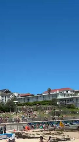 The video captures a beach scene on a sunny day under a clear blue sky. A crowd of people is visible on a grassy area and on the beach. Some are sunbathing, while others are walking around. Buildings are visible in the background, including some with red tile roofs. There is also a yellow warning sign near the beach. The tide appears to be out, with some seaweed or algae visible on the sand in the foreground.