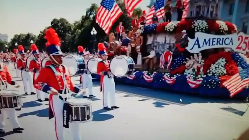 A marching band is parading down a street. The band members are wearing red uniforms with blue and white accents, and tall, feathered hats. They are all playing drums. To the right of the band is a large float decorated with red, white, and blue flowers. The float has the word "AMERICA" written on it in large letters. American flags are flying on the float and in the background. The environment appears to be outdoors, possibly on a sunny day, with trees visible in the background.