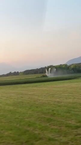 The video shows a passing view of a rural landscape. In the foreground, a dirt road is visible on the left. Beyond that, a green field stretches out, with a cornfield visible in the middle ground. An agricultural irrigation sprinkler is actively spraying water in the cornfield. In the distance, trees and hills form the horizon, under a pale sky transitioning from blue to a soft orange hue, suggesting either dawn or dusk. The camera appears to be moving, possibly from a vehicle.