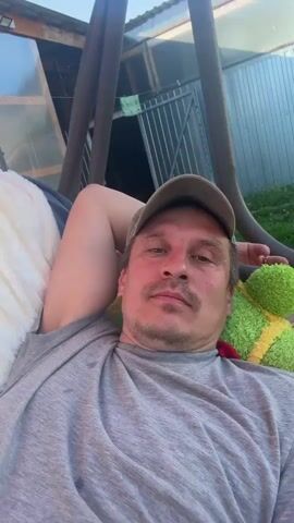 A middle-aged man is lounging in a swing outdoors, facing the camera. He is wearing a grey t-shirt and a baseball cap. He is smiling. Behind him is a white blanket, a green stuffed animal, a greenhouse, and a fence. The sun is shining. Birds chirp in the background.
