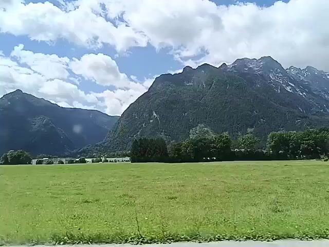 The video shows a scenic view of mountains and a grassy field. The sky is partly cloudy with blue sky visible. There is a treeline in the middle ground and a house on the right side of the frame. In the foreground, there is a tripod with a reflective object on top, which appears to be a camera or some other recording device. The camera is panning from left to right, capturing the landscape. The footage is shaky, indicating it was likely recorded while moving.