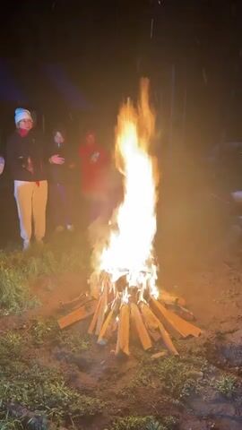 At night, a bonfire blazes brightly in an outdoor setting. The fire is built on the ground from logs arranged in a conical shape. Five people stand near the fire. The person closest to the camera wears a black jacket, white pants, and a light blue beanie. Another person in the group wears a red jacket. The other three people are further away. The ground around the fire is covered with grass. In the background, there is a dark structure with a blue tarp visible. The lighting is dim, with the fire providing the primary source of illumination.