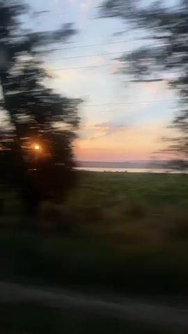 This is a video of a sunset over a body of water, viewed from a moving vehicle. The sun is low on the horizon, casting a warm glow across the sky. There are some clouds scattered in the sky, and rays of light are breaking through them. In the foreground, there is a field or meadow, and in the distance, there is a body of water. The video is somewhat blurry due to the movement of the vehicle, but the overall scene is serene and beautiful.
