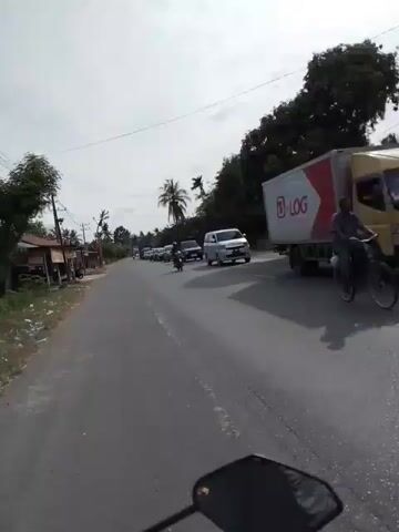 A motorcyclist's point-of-view shot shows a moderately busy asphalt road with several cars and trucks traveling in the same direction. The road appears to be in a rural or semi-rural area, with vegetation and some small buildings visible on the left side. The weather is overcast, and the sun is not visible. The traffic seems to be moving slowly, suggesting a potential congestion or an upcoming intersection. The camera focuses on the road ahead and the vehicles in front, with the handlebars and mirrors of the motorcycle visible at the bottom of the frame.
