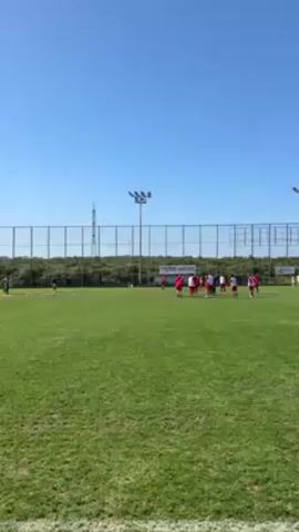 The video depicts a soccer team on a green field during what appears to be a training session. The scene takes place outdoors on a bright day with a clear blue sky. The field is surrounded by a wire fence, and a few light posts are visible along the perimeter.  A group of players, wearing red and white uniforms, is gathered in the center of the field. They appear to be either listening to instructions or preparing for a drill. Several soccer balls are scattered on the field near the group. In the background, beyond the fence, there's some greenery, possibly trees or bushes.  The camera is relatively static, capturing the scene from a distance, allowing a broad view of the training area and the players involved.