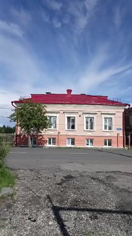 The video shows a street view with two old buildings on the left side of the frame. The first building is painted light pink and has a red roof. It has several windows on the upper floor and smaller windows on the lower floor. A tree is planted in front of this building. Next to it, to the right, is a second building made of brick, with a more dilapidated appearance. It also has windows on multiple floors, some with shutters. A pink metal gate is visible in front of this building. Further to the right, beyond the brick building, another structure and some trees can be seen. A street runs in front of the buildings, with a paved surface that appears worn and has puddles. A concrete utility pole with wires stands near the right side of the frame, and cars are parked along the side of the street. The sky is blue with some white clouds, and the sun is shining. The camera appears to be handheld and pans slightly to the right.