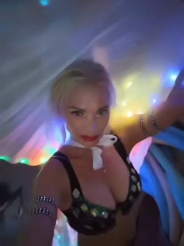 The video features a woman with blonde hair and red lipstick, dressed in a sparkly bra and armbands, dancing in a room illuminated by colorful lights. She is holding a sheer, flowing fabric that is also lit by the lights, creating a mesmerizing effect. The woman appears to be in good spirits, smiling and engaging with the camera. The room's decor is dimly lit, with the focus primarily on the dancer and the vibrant lights. The overall atmosphere suggests a festive or performance-oriented setting, possibly a stage or a decorated room for an event. There are no discernible objects or text that provide further context about the specific activity or location beyond the visual cues of a performance.