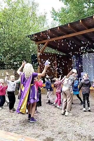 A group of children and two adults are gathered in an outdoor area, likely a park or yard, under a wooden structure. A large amount of soap bubbles fills the air around them, creating a festive atmosphere. The children are of various ages, possibly ranging from preschool to elementary school, and are wearing different casual clothes. One adult, positioned towards the left side of the frame, is wearing a long blonde wig and a purple and yellow cape. They appear to be interacting with the children, possibly as a performer or entertainer. The other adult is wearing a white cap and beige pants, and is watching the children. The wooden structure has a roof and decorative lattice, and behind it is a clear plastic sheet or curtain. The ground is covered in gravel. Trees are visible in the background, suggesting the area is surrounded by greenery. The children and adults seem to be enjoying the bubbles, reaching out to catch them. The lighting is soft and natural, indicating an overcast day.