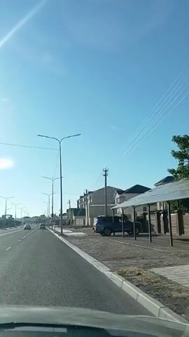 This is a vertical video taken from a car driving down a paved road on a clear, sunny day. The sky is a vibrant blue with a bright sun casting lens flare on the left side of the frame. The road is asphalt with white lane markings and a concrete curb.

Along the right side of the road, there are several modern streetlights with two lamps each, spaced at intervals. Beyond the streetlights, a sidewalk or paved area is visible, and then a row of contemporary houses and buildings. These structures are mostly light-colored with dark roofs, and some appear to be two or three stories high. One building has a green roof. There are also utility poles with wires running overhead.

A dark blue SUV is parked on the right side of the road, partially under a covered structure. Other cars are visible further down the road in the distance, traveling in the same direction as the camera. There are also some trees and landscaping elements.

A triangular road sign with a red border and a symbol inside is visible on a pole near the roadside. Another pole has a circular device mounted on it, possibly related to utility or communication equipment. There are no people clearly visible in the video.

The overall impression is a drive through a developing suburban or exurban area, likely in the morning or early afternoon given the strong sunlight and clear skies. The environment appears tidy and modern.