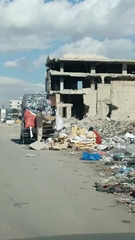 The video depicts a man loading items onto the back of a truck, in a setting marked by destruction and debris. The truck is already partially filled with large bags, some appearing to be filled with plastic bottles. The man is dressed in neutral-colored clothing. He is picking up cardboard and other objects from a pile on the ground and placing them onto the truck bed. The surrounding environment is strewn with trash and debris, suggesting an area affected by conflict or severe neglect. In the background, there is a partially destroyed building. The top floors are damaged, with exposed support structures and gaping holes in the facade. The sky is partly cloudy, and the overall scene conveys a sense of devastation and hardship.