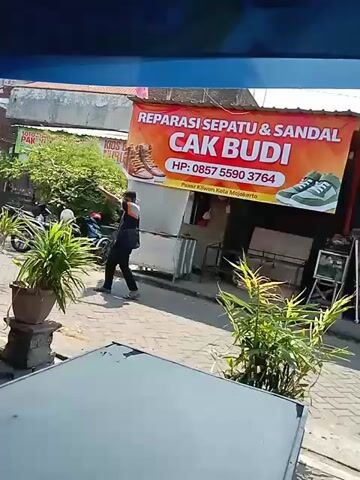 The video shows a man walking past a shop called "Reparasi Sepatu & Sandal Cak Budi" which translates to "Shoe & Sandal Repair Cak Budi". The shop has a banner displaying the name of the shop, images of shoes and sandals, and a phone number: 0857 5590 3764. The man is wearing a dark shirt and pants and appears to be walking away from the shop. The shop is located on a street with other businesses and a motorcycle parked nearby. The environment appears to be a marketplace or a commercial area. The overall scene suggests a typical street view in Indonesia.