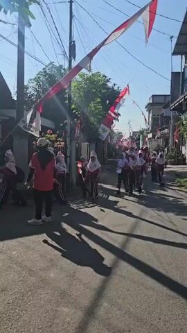 A group of people, primarily young students in uniform, are walking down a residential street. The street is lined with buildings and decorated with Indonesian flags strung across the road. The students appear to be participating in some sort of parade or procession. One person in a red shirt and hijab is standing on the left side of the road, facing the students. The sun is shining, casting long shadows on the asphalt.