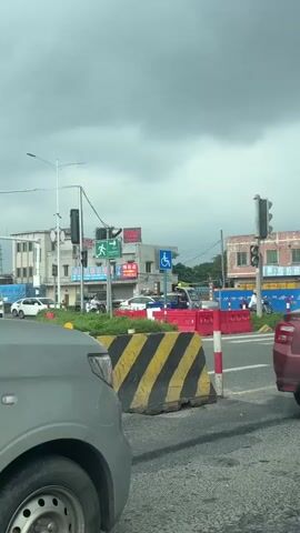 The video shows a street view under a cloudy sky. In the foreground, the hood and wheel of a gray car are visible. To the right, a portion of a red car is also in view. In the middle ground, there is an intersection with traffic lights. Several vehicles, including white cars and a red car, are stopped at the intersection. Construction barriers, some with yellow and black diagonal stripes and others red, are placed around the intersection. Signs are visible, including a pedestrian crossing sign with an arrow and a blue sign with a wheelchair symbol indicating accessibility. Buildings line the street in the background. A large orange truck is visible in the far left background, followed by a white truck. The overall scene appears to be a typical urban traffic intersection with ongoing construction.