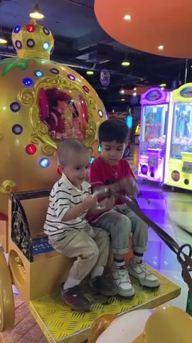The video shows two young children sitting on a horse-drawn carriage ride in an arcade. The older child is wearing a red shirt and grey pants with sneakers, while the younger child wears a striped shirt and tan pants. The carriage is gold and white with colorful lights and a picture of a prince and princess. The carriage ride is surrounded by claw machines and other arcade games. The arcade has a bright orange ceiling with various hanging decorations. Other people can be seen walking around in the background. The children are interacting with the reins of the horses, pretending to drive the carriage. The camera moves around the carriage, showing the details of the ride and the arcade environment.