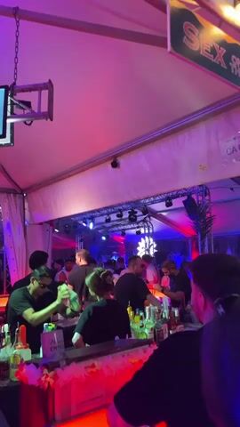 This media file depicts a lively outdoor event, possibly a festival or beach party, under a large white tent. The scene is illuminated by vibrant purple and pink lighting, creating a festive and energetic atmosphere.

The primary subjects are a crowd of people at a bar. Several individuals are visible at the bar, some appearing to be staff serving drinks, while others are patrons. The bar itself is stocked with numerous bottles of alcohol and is decorated with colorful leis. In the background, more people are mingling and enjoying the event.

The activity taking place is a social gathering centered around a bar, likely involving drinking and socializing. The mood appears to be upbeat and celebratory, as suggested by the lighting and the presence of a crowd.

Notable details include several digital menu boards hanging above the bar, displaying drink options and prices. One sign is partially visible with the word "SEX" on it, suggesting a playful or themed aspect to the event. Another sign reads "BEACH," indicating the proximity to a coastal area. The lighting suggests it is nighttime or evening. The location is identified as Kiel, Germany, which aligns with the "BEACH" sign and the presence of what appears to be a harbor or marina in the background, with boats visible.

Visible text includes:
- A sign with "SEX"
- A sign with "BEACH"
- Text on digital menu boards, some of which appears to be in German, showing drink names and prices like "5,00€" and "15,00€". One board has a heading that looks like "SOMMER SPECIAL". Another seems to list "COCKTAILS" and "LONGDRINKS".
- Text on a sign below the bar appears to read "Wimpel Erfurt" and "5,00€".