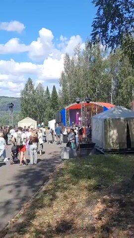 A group of people are gathered outdoors at what appears to be a festival or event. There is a stage set up with lights and speakers. Several people are walking along a paved path, some are standing and talking, and some are seated. The weather is sunny with scattered clouds. There are trees and rolling hills in the background.