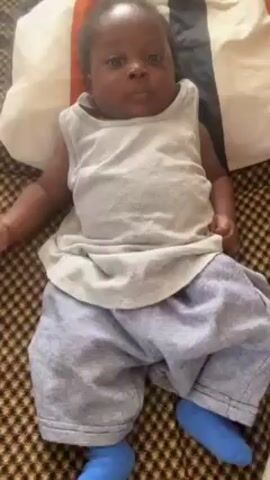 A baby lies on their back on a patterned fabric surface. They are wearing a grey tank top, light blue pants, and blue socks. A pillow with a striped cover is behind the baby's head. The baby's expression changes from neutral to crying.