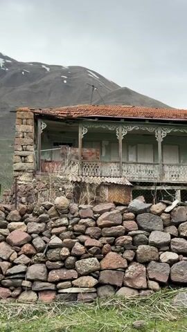 The camera pans from left to right across the facade of a dilapidated two-story house perched atop a stone wall. The wall is made of large, irregular stones in shades of grey, brown, and red, and a small patch of green grass grows at its base. The house, painted a faded light green, features a distinctive wooden porch with intricate fretwork. The porch doors and windows are mostly closed with white shutters. The roof is tiled in a reddish-brown color, with visible signs of wear and damage.  Behind the house, a mountain with a dusting of snow on its peak is visible under a gray, overcast sky. A bare tree stands to the right of the house, adding to the somber and neglected atmosphere. The scene evokes a sense of abandonment and decay.