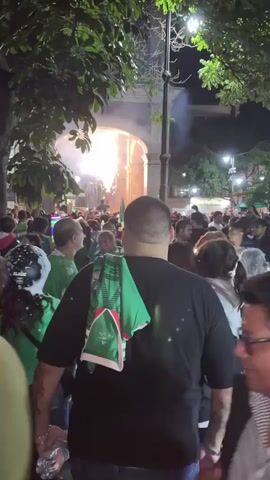 The video shows a crowded outdoor scene at night. The crowd is densely packed, with many people wearing green shirts. There are trees and a large archway structure in the background. Fireworks are visible in the air, emitting sparks and smoke. Streetlights and artificial lighting illuminate the area. Some individuals are holding what appear to be flags or scarves, also in green. The overall atmosphere suggests a celebration or gathering. The camera is moving, and the focus shifts between different individuals and the general crowd. Some people are looking towards the fireworks, while others are engaged in conversation or looking in different directions. The lighting conditions create a somewhat hazy and vibrant effect.