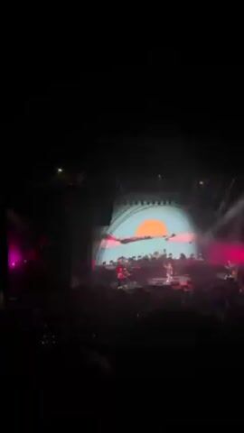 A crowd of people is attending a concert at night. The stage features a large screen showing the performer and a complex structure with lights. Some people in the crowd are raising their hands. The atmosphere seems energetic and lively.