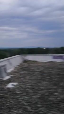 The video shows a person walking across a flat roof. The ground is rough, black, and possibly pebbly. A low white wall runs along the edge of the roof, with purple graffiti visible. In the background, there are trees and some buildings, with a cloudy sky above. As the person walks, they initially wear a white shirt, then appear to change into a blue shirt and carry a black backpack. They are holding a phone or similar device and seem to be taking pictures or video. The camera movement is shaky, likely due to the person filming while walking.