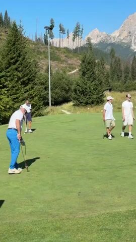 Four people are on a golf course. Two men are on the putting green, one is addressing the ball as if to putt. The other is standing behind him. Two young men are walking on the green away from the camera, one is carrying a golf club. In the background are trees and mountains under a clear blue sky.