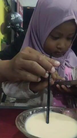 The video shows a young child wearing a purple hijab and a gray sweater over a pink shirt. The child is sitting at a table with a clear bowl of white liquid and a black straw. An adult's hands are visible, guiding the straw to the child's mouth and holding a smartphone. The child is looking at the smartphone while drinking from the straw. The background is dark, with some blurry lights and what appears to be a motorcycle. The video is shaky and the lighting is dim, suggesting it was filmed in a low-light environment, possibly at night. The overall scene suggests a casual, possibly outdoor or roadside setting, where the child is being entertained with a phone while having a drink.