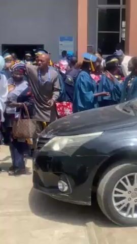 The video shows a group of people gathered outside a building, some of whom appear to be celebrating a graduation. A dark-colored car is parked at the edge of the crowd, and several people are standing near it. Some individuals are wearing graduation gowns and caps, while others are dressed in traditional African attire. There are also people in business casual clothing. The building behind the group has signage indicating TETfund and mentions "Normal Intervention." Another sign shows the number 9964. There are multiple cars in the scene, including a white car with a "PLATEAU" license plate visible. The atmosphere seems lively and celebratory.