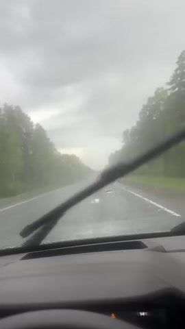 The video shows the view from inside a car driving on a wet road. It is raining heavily and the windshield wipers are on. The dashboard is visible, displaying information such as speed (82 km/h), distance traveled, fuel consumption, and other data. The road ahead is wet and there are trees on both sides. Another car is visible in the distance. The sky is overcast and gray.