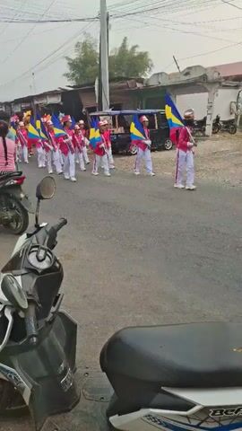 The video shows a street scene with a marching band. Several people, mostly young individuals, are walking in formation, wearing matching pink and white uniforms. They are carrying flags with yellow, blue, and pink colors. Some of the individuals are holding what appear to be drumsticks. The street is paved and lined with buildings and what look like houses or small shops on either side. There are also several motorcycles parked and being ridden in the video, suggesting a public event or parade. The weather appears to be overcast. The camera is positioned at a low angle, capturing the marching band from the side and slightly in front.