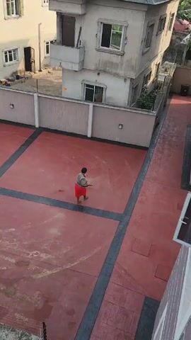 From an elevated perspective, the video shows a woman sweeping an outdoor concrete area in what appears to be a residential setting. The woman is wearing a gray shirt and a red skirt, and sandals. She is using a traditional broom made of twigs or straw. The concrete surface is red with dark gray borders delineating square sections. The area is surrounded by buildings with neutral-colored exteriors and a short perimeter wall. There are also some outdoor light fixtures mounted on the walls. The lighting suggests it's daytime. The woman is focused on sweeping the area, moving debris in a circular pattern. There are apparent marks or stains on the concrete.