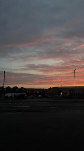 The video shows a time-lapse of a sunset over a commercial area. In the foreground, there is a dark asphalt road. In the background, there are several buildings, including what appears to be a supermarket with a yellow logo. A street lamp stands to the right of the buildings. Cars are visible on the road in front of the buildings. The sky is filled with dramatic clouds that transition from gray to shades of pink and orange as the sun sets.