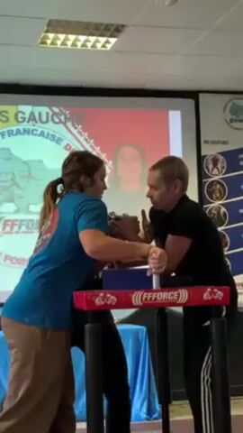 In an indoor setting, two individuals are engaged in an arm wrestling match. A man in a black shirt and black pants with white stripes is facing a woman in a blue shirt and brown pants. They are both gripping hands on an arm wrestling table marked with "FFFORCE". A referee stands to the side, observing the match. Behind them, a projector displays a poster with text in French, including the words "LAS GAUCHE" and "FEDERATION FRANCAISE DE FORCE." The room appears to be a conference or training space, with fluorescent lighting and additional posters visible on the wall. The participants are focused on the competition, displaying tense facial expressions.