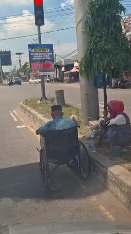 A man in a wheelchair faces away from the camera, positioned on the edge of a road next to a sidewalk. He is wearing a blue patterned shirt and a dark cap. To his right, a woman wearing a red headscarf and a patterned skirt is seated on the ground under a tree, also facing away from the camera. A small green bucket and a water bottle are visible near her. In the background, a traffic light is red, and vehicles are stopped at an intersection. A sign with red lettering pointing to the right is visible behind the man. The environment appears to be an urban or semi-urban area with buildings and street infrastructure visible.