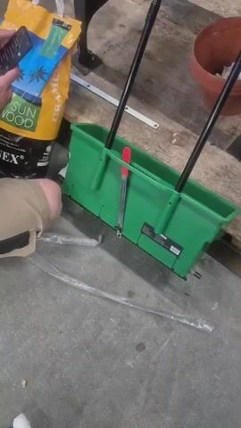 A person is sitting on a concrete floor. In front of them is a green spreader with two black handles. To the left of the spreader is a yellow bag of gardening supplies with a picture of palm trees on it. The person is holding a smartphone. To the right of the spreader is a brown pot.