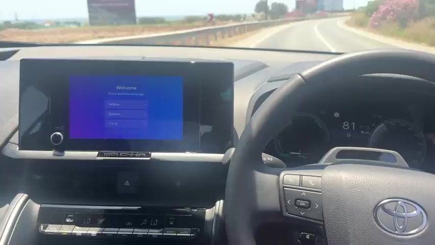 This video provides an interior view of a car driving along a coastal road in Timi Community, Cyprus. The perspective is from the driver's seat, showing the dashboard, steering wheel, and the road ahead through the windshield.

The primary subjects are the car's interior elements. The dashboard features a prominent infotainment screen displaying a "Welcome" message with a list of languages, including "English," "Español," "Français," "Deutsch," and "Dansk." To the right of the screen is the steering wheel with visible controls and the car's digital instrument cluster. The cluster shows a speed of 81 (likely km/h given the location context), a "D" for drive mode, and a power/charge indicator. The overall car model is identifiable as a Toyota C-HR from the badging on the dashboard.

The activity captured is the car in motion, driving on a paved road. The road curves gently to the right, bordered by a guardrail on the left. Beyond the guardrail, the Mediterranean Sea is visible, with a coastline dotted with vegetation and some buildings. In the distance, billboards and utility poles are visible along the roadside.

The time of day appears to be daytime, with bright sunlight casting shadows and indicating clear weather. The clear sky and bright lighting suggest a pleasant day for driving. The location cues, such as the sea, coastal vegetation, and road signage (a curved arrow warning sign), along with the explicit mention of Timi Community, Cyprus, establish the setting.

There are no people visible within the car's interior, and the focus is on the driving experience and the car's technology. The overall mood is neutral, reflecting the functional aspect of recording a drive.

Notable visible text includes the car's model name "TOYOTA C-HR," the language options on the infotainment screen, and the speed displayed on the instrument cluster. There is also a sign visible in the distance, but the text is too small to read.