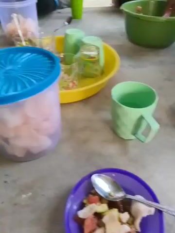 The video shows a group of people sitting on the ground, seemingly having a meal or a snack. In the foreground, there is a purple plate with food, and a spoon is interacting with it. To the right, there is a large green pitcher, and next to it, a clear plastic container with a blue lid, containing what appears to be light-colored food items. Further to the right, a yellow bowl holds several green cups. In the background, more people are visible, also sitting on the ground. One person is wearing a hijab and a beige top with dark stripes, and appears to be eating something. Another person is wearing a red top. The environment appears to be outdoors, with dirt and some greenery visible in the background. The overall impression is a casual gathering.