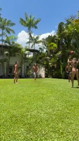 The video shows a sunny outdoor scene where four people are playing with a ball on a large, green lawn. Three of the individuals appear to be men, and one is a woman. All are wearing minimal swimwear. The group is spread out across the lawn, tossing the ball to one another. Behind them, there is a large white building with windows and multiple palm trees. Dense tropical vegetation frames the lawn on the right side of the video. The sky is blue with scattered clouds.
