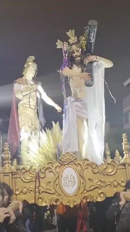 The video shows a religious procession at night. In the center, a statue of Jesus Christ, crowned with thorns and draped in a white cloth, carries a large cross on his right shoulder. To his left stands a statue of a Roman soldier in armor, holding a whip. The statues are adorned with purple and gold streamers. They are mounted on a golden float decorated with ornate carvings and the monogram "JHS." The float is being carried by several people, whose arms and shoulders are visible supporting the weight. In the foreground, the faces of several women are visible, looking towards the float. The background is dark, suggesting nighttime, with some faint lights and building structures visible. The scene appears to be a solemn religious event, likely part of Holy Week celebrations.