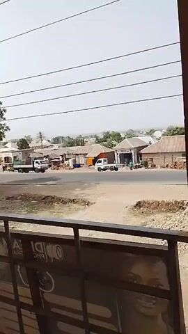 The video shows a street scene from an elevated perspective, likely a balcony. A paved road runs horizontally across the frame with various buildings lining the road, mostly single-story structures with corrugated metal roofs. Several vehicles are visible, including a few small trucks parked along the road and a motorcycle driving on it. Some people are standing near the buildings. Overhead, power lines stretch across the sky. In the foreground, a metal railing obscures the bottom portion of the view, partially hiding a large advertisement featuring a photograph of a woman's face. The overall impression is of a small town in a developing area. The lighting suggests a bright day, but there appears to be some haze or dust in the air.